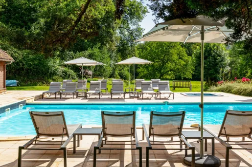 Outdoor pool surrounding by reclining chairs and umbrellas, with large trees in the background