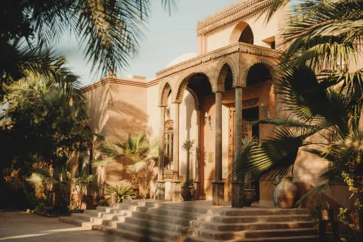 The main exterior entrance of the Al Moudira Hotel in Luxor, Egypt with traditional architecture, featuring hand-carved wooden arches, clay-colored mudbrick structures, and surrounding palm trees.