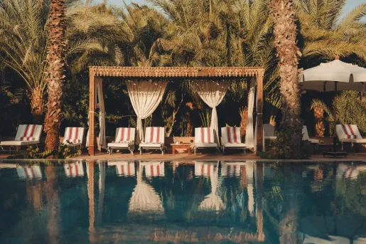 The outdoor swimming pool area at the Al Moudira Hotel in Luxor, Egypt features a large grove of mature date palms, striped lounge chairs, and a hand-carved wooden cabana structure.