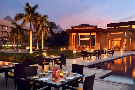 The outdoor dining terrace at the historic Marriott Mena House, Cairo in Giza, Egypt at twilight with the The Great Pyramids of Giza in the background, providing an iconic backdrop for guests dining by the water.