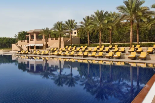 The main swimming pool area at the Marriott Mena House, Cairo in Giza, Egypt features rows of sun loungers with bright yellow cushions that line the multi-tiered stone deck with palm trees overlooking the water.