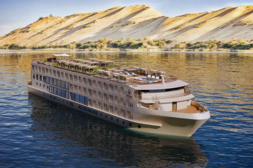 Luxury river cruise ship sailing on the Nile River, featuring multiple passenger decks, outdoor lounge areas, and a rooftop terrace. Golden desert hills rise along the riverbank in the background.