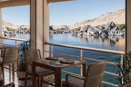 Open-air dining area aboard a luxury Nile River cruise ship, featuring wooden tables and woven chairs positioned beside large open windows. The view overlooks calm river waters, granite rock formations, and golden desert hills.