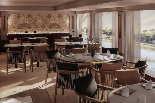 Elegant restaurant aboard a luxury river cruise ship, with round and rectangular tables, upholstered chairs, decorative wall panels, and floor-to-ceiling windows offering views of the Nile River and shoreline.
