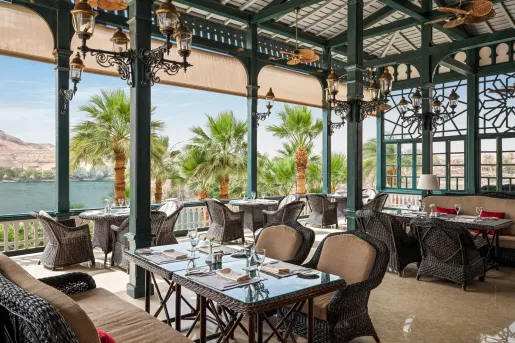 Elegant, open-air terrace restaurant with green wooden pillars, wicker dining tables, and plush seating overlooking the Nile River at the Old Cataract, Aswan in Egypt. 