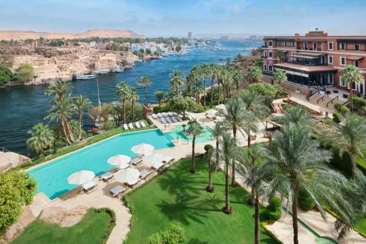 A large infinity-style swimming pool lined with white sun loungers and umbrellas at the Old Cataract Aswan, showcasing its landscaped grounds overlooking the Nile River.