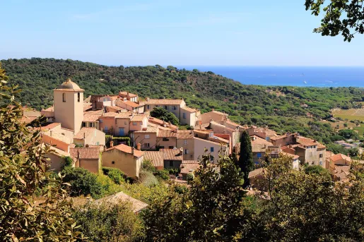 View of a Mediterranean hillside village with terracotta rooftops and a church bell tower, surrounded by green countryside and overlooking the sea in the distance.