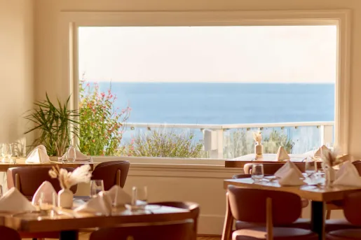 Interior of a restaurant in British Columbia, Canada, with a stunning view of the ocean through a large window. The room is lit with natural sunlight streaming in, casting soft shadows on the wooden floor. Several round wooden tables are set with white tablecloths, folded napkins, and glassware, ready for diners.