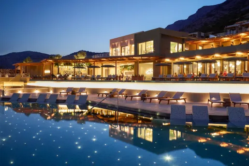 Luxury resort pool area surrounded by tropical landscaping, lounge chairs, and private cabanas. Outdoor dining seating sits beside the pool, with views toward the ocean through the trees in the background.