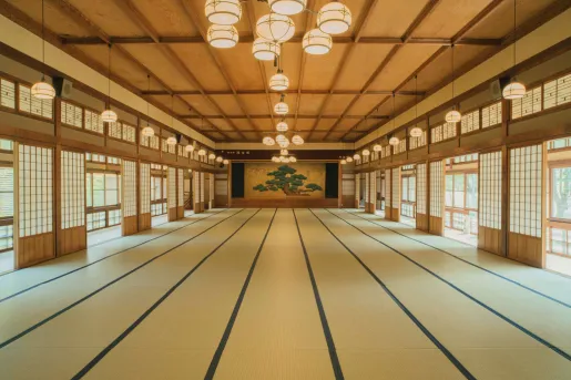 Spacious tatami hall at Ochiairo Hotel in Japan, showcasing traditional Japanese architecture with shoji screens, wood-paneled ceilings, hanging lanterns, and a decorative pine tree mural at the far end of the room.