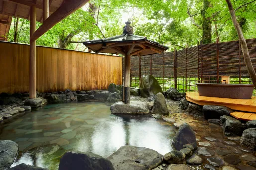 Outdoor onsen at Ochiairo Hotel in Japan, featuring a stone-lined hot spring pool surrounded by natural rock elements, bamboo screens, and lush forest greenery. A traditional wooden shelter and soaking tub create a serene setting immersed in the Japanese countryside.