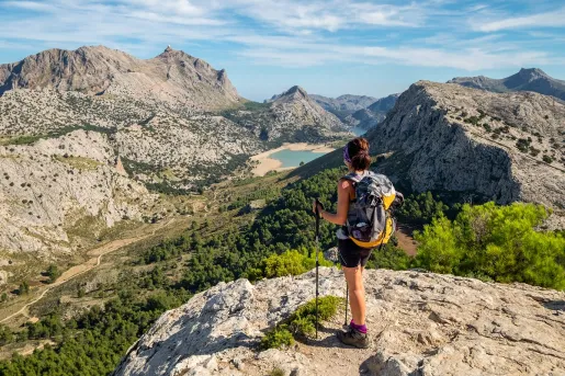 Woman standing on top of a cliff holding two hiking poles, looking down towards tall mountains