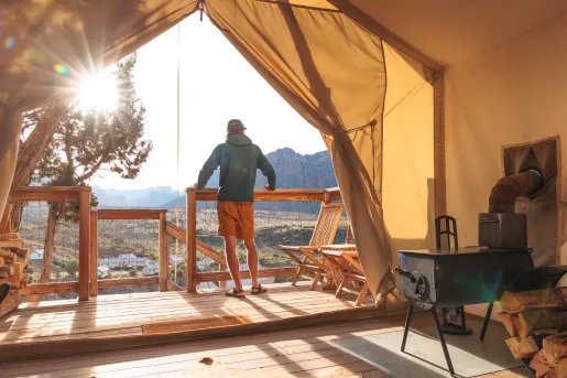View from inside a safari-style glamping tent overlooking a desert valley and distant red rock mountains. A wooden deck with chairs sits outside, while sunlight streams through the open tent entrance.