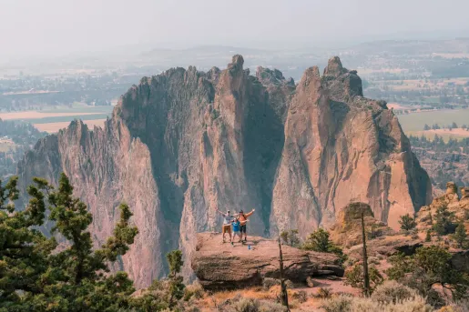Visitors stand on a rocky overlook with arms raised, surrounded by dramatic rock pinnacles and sheer canyon walls at Smith Rock State Park, with the high desert landscape stretching into the distance.