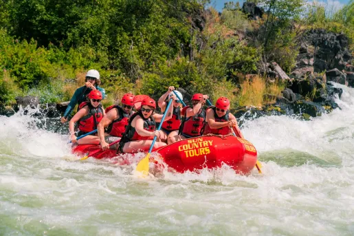 Group of rafters wearing helmets and life jackets paddle a red raft through whitewater rapids on a river near Smith Rock, surrounded by rocky banks and green vegetation.