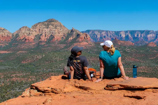 Two travelers sit on a rocky overlook taking in panoramic views of Sedona’s red rock mountains and valleys.