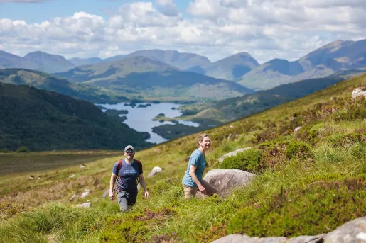 Two people ascending a grass and moss-covered hill, with a lake and mountains in the distance