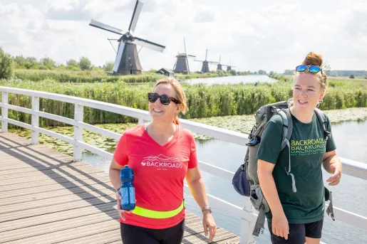 Two women smiling while walking on a bridge, with large windmills in the background