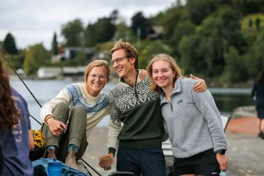 One man and two women with arms around their shoulders, smiling with a large lake behind them