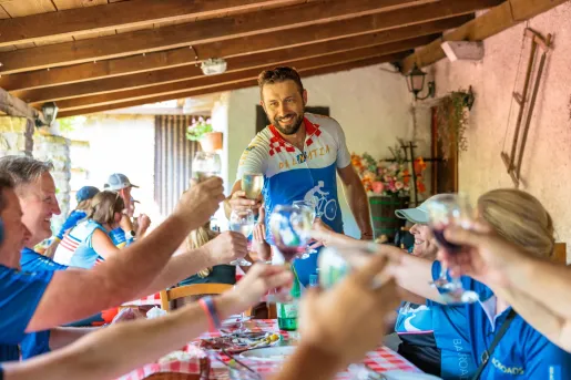 Cyclists gather around a long table at a rustic outdoor restaurant, raising glasses in a toast beneath a covered terrace with wooden beams and checkered tablecloths.