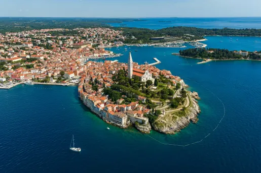 Aerial view of the historic coastal town of Rovinj, Croatia, with terracotta rooftops clustered on a peninsula extending into the Adriatic Sea. A prominent church bell tower overlooks the harbor, marinas, and surrounding blue waters.