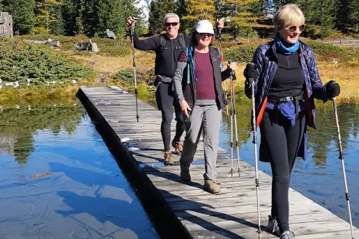 Three hikers use trekking poles to cross a wooden boardwalk spanning a shallow alpine wetland. Evergreen trees, patches of snow, and distant mountain peaks surround the landscape beneath a clear blue sky.