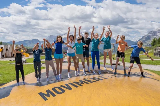 Group of children jump on a large outdoor inflatable pillow at a mountain activity park, with alpine scenery, grassy meadows, and distant mountain peaks under a sky filled with fluffy clouds.
