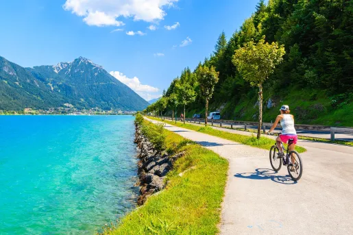 Woman biking on a road next to a lake, with a hill covered in trees to the right