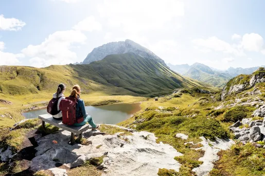 Two women sitting on a bench, looking out towards a valley with a small lake and hills