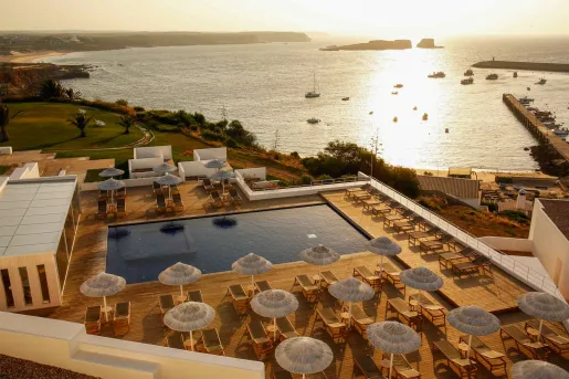 The pool deck area at Memmo Baleeira Hotel in Sagres, Portugal, features a modern, minimalist design and white umbrellas with panoramic views of the Atlantic Ocean. 