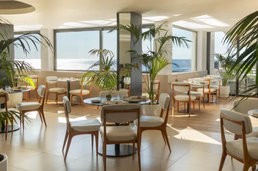 The modern dining area of the Memmo Baleeira Hotel featuring floor-to-ceiling sea views and minimalist design with white cushioned dining chairs and palm tree plants.