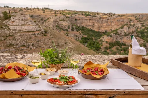 An outdoor table spread features wine glasses, fresh strawberries, bruschetta, and olives set on a white tablecloth.