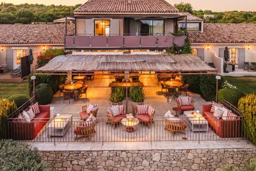 A multi-level stone and stucco villa features an outdoor restaurant and lounge setup with several round tables with red striped chairs at Hôtel La Bastide Saint Georges.