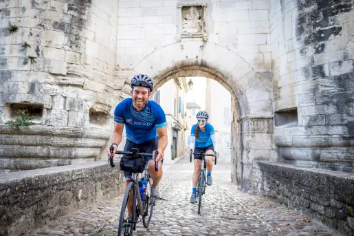 Two cyclists ride on a cobblestone street through a historic stone gateway. Tall medieval walls and an arched passage frame the narrow street in a preserved old town setting.