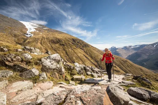Woman with hiking poles, walking on larger boulders next to a mountain