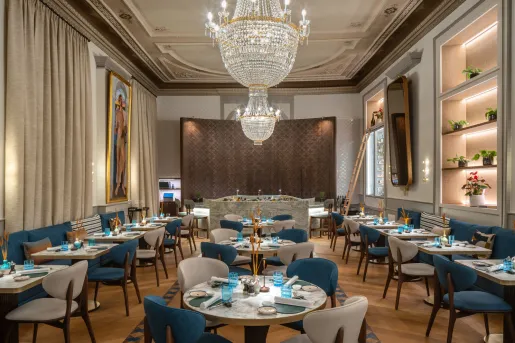 The elegant dining space featuring a tiered crystal chandelier, curved plush velvet seating in deep blue, and warm wooden herringbone floor at the Grand Hotel des Bains Kempinski located in St. Moritz, Switzerland.
