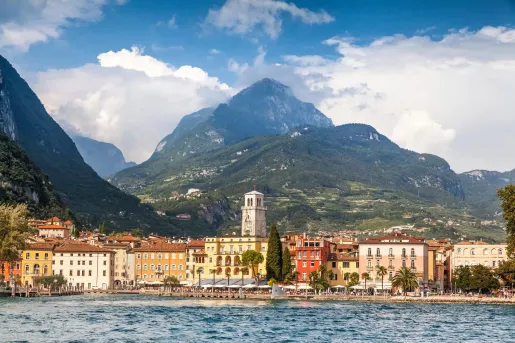 View across a lakeside town with colorful waterfront buildings and a historic clock tower, backed by forested mountains and dramatic peaks rising above the shoreline.