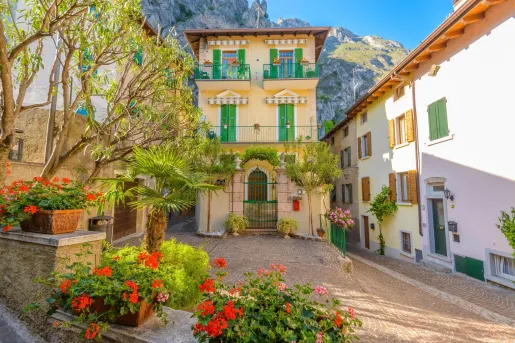 Charming village courtyard featuring colorful flowers, potted plants, and a historic building with green shutters and balconies. Steep rocky mountains rise behind the village beneath a clear blue sky.
