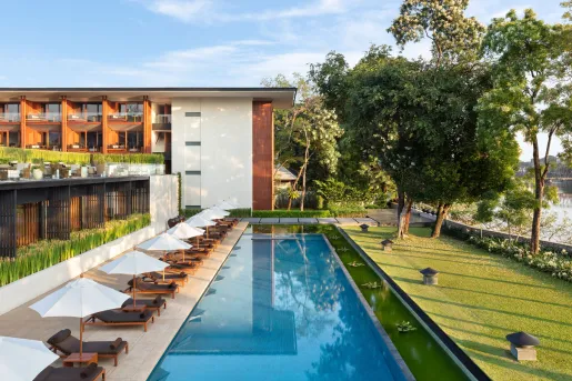 White and brown building with a long, outdoor pool surrounded by chairs and grass fields