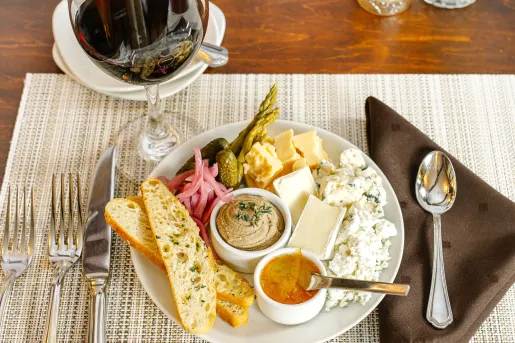 A snack board featuring a variety of curated cheeses, pickled onions, cornichons, a small bowl of pâté or spread, garnished with toasted bread slices and a citrus marmalade.