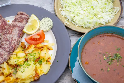 A meal featuring a grilled steak served with bratkartoffeln (German-style pan-fried potatoes), a cabbage salad and a bowl of soup.