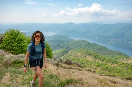 A woman wearing sunglasses hikes up a hill with Lake Como in the distance.