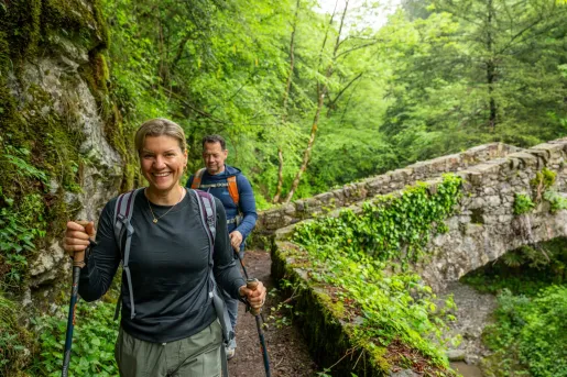 A hiker smiling hiker and her friend hike across a cobblestone bridge in a lush Italian forest.