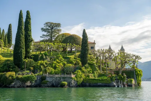 A beautiful collection of trees and other plants surround a building that sits on the shore of Lake Como.