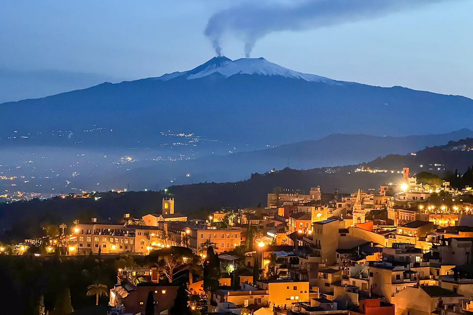 Nighttime shot of Italian hillside town, smoking volcanoes in distance.