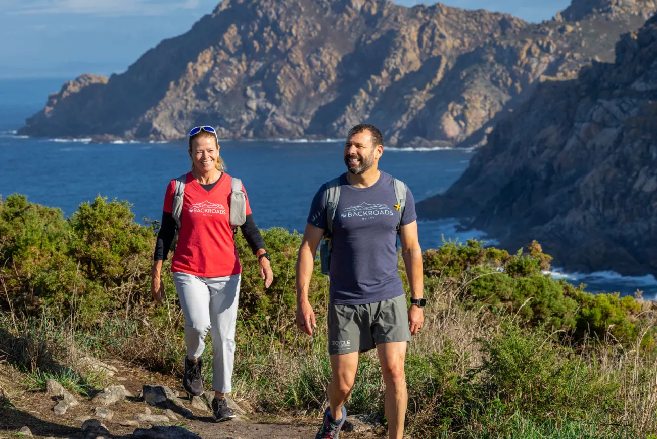 Man and woman smiling while walking on a dirt and stone path and the ocean in the background