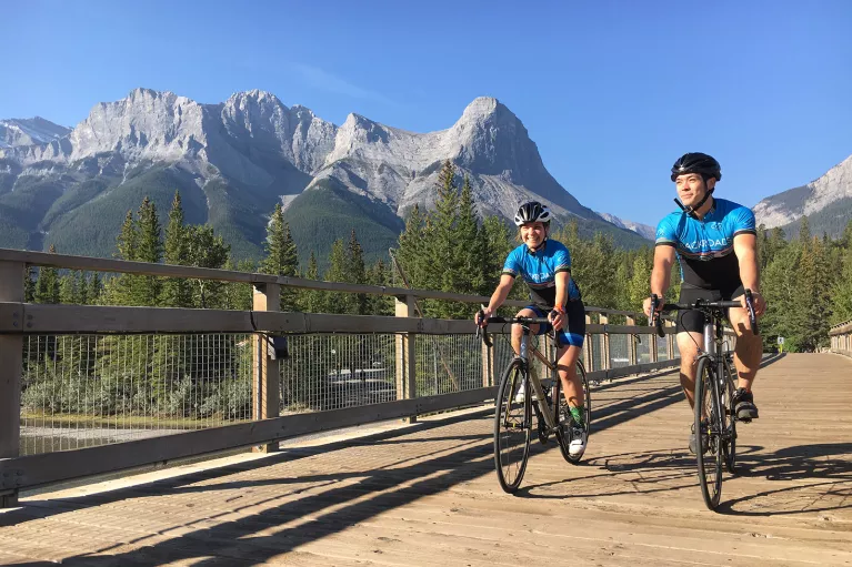 Two guests cycling on wooden path, Rockies in distance.