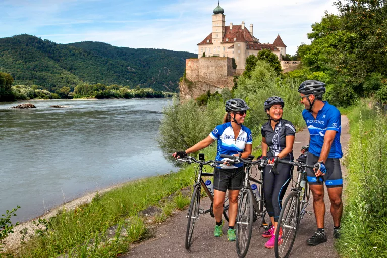 Three bikers walking their bikes on a path along the Danube River.