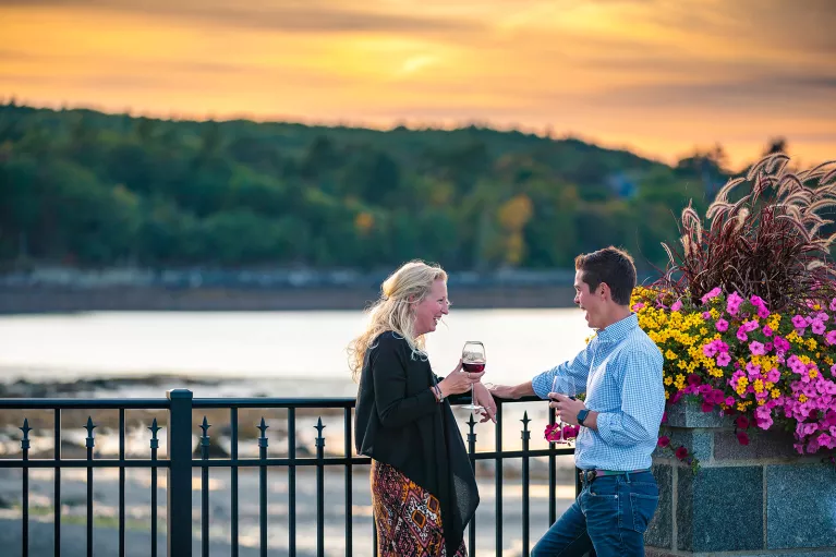 Two guests chatting, wine glasses in hand, sunset and beach in background.