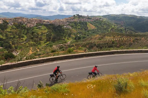 Two guests cycling down road, Medit. hilltop town in distance.